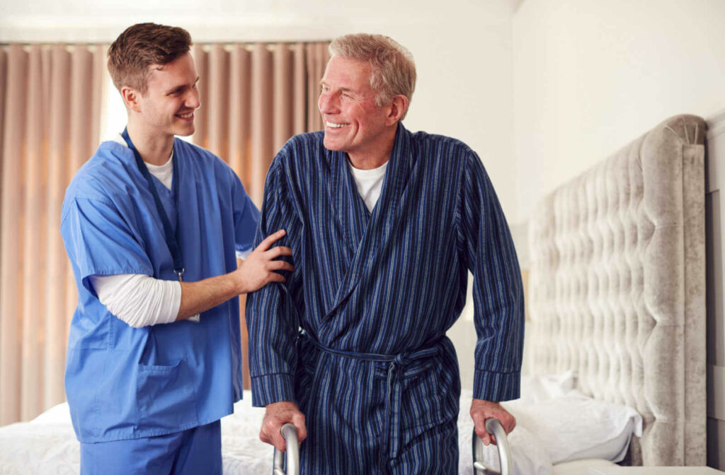 A male nursing assistant in a blue scrub suit is assisting an elderly man in walking. The old man is wearing a blue stripe robe and both hands on his steel cane walker.