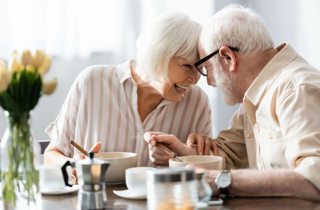 A happy white-haired senior couple having breakfast on the table with a bowl of cereal and cups of coffee.