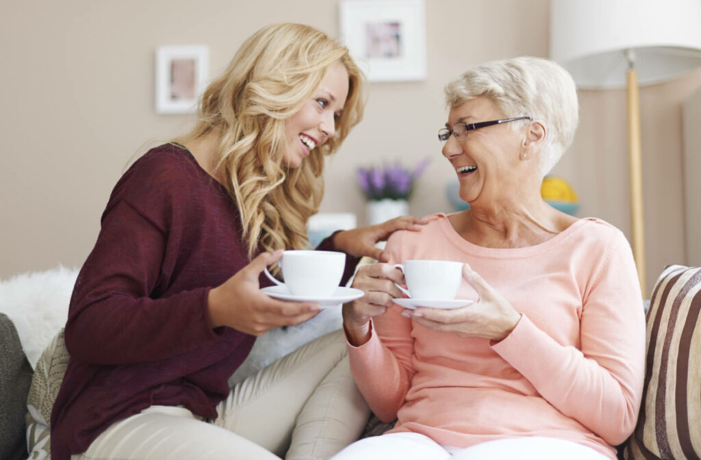 A grown daughter and her elderly mother sit on a living room couch, chatting and enjoying a cup of coffee together.
