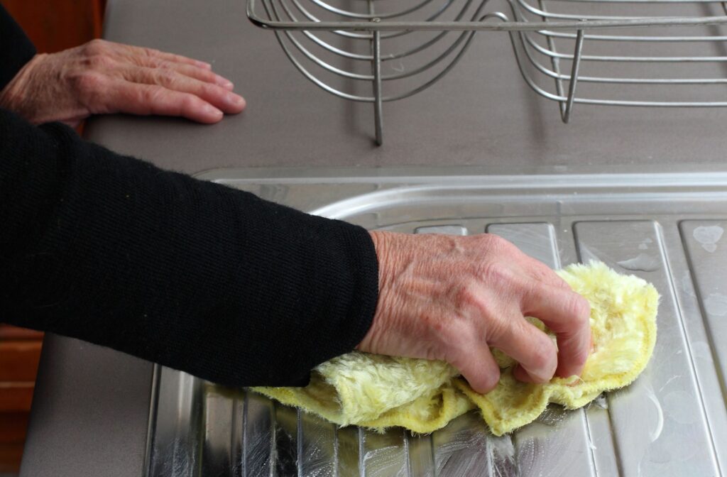 A hand holds a cloth, cleaning the stainless steel kitchen sink counter.